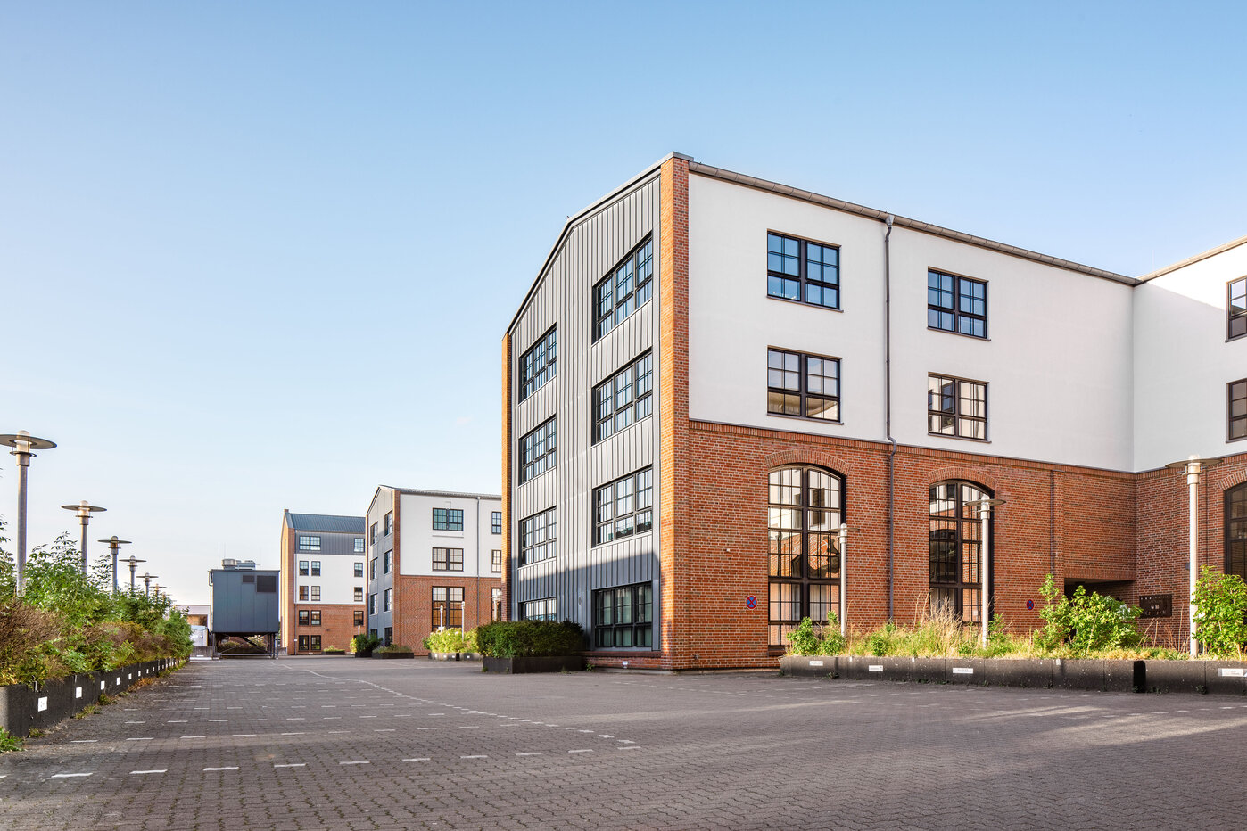 Moderne Wohngebäude im urbanen Raum Die Abbildung zeigt moderne Gebäude in einer urbanen Umgebung. Im Vordergrund ist ein gepflasterter Platz, der Platz für Fahrzeuge bietet. Die Architektur ist zeitgenössisch mit großen Fenstern und klaren Linien, umgeben von einigen Pflanzen und Straßenlaternen.