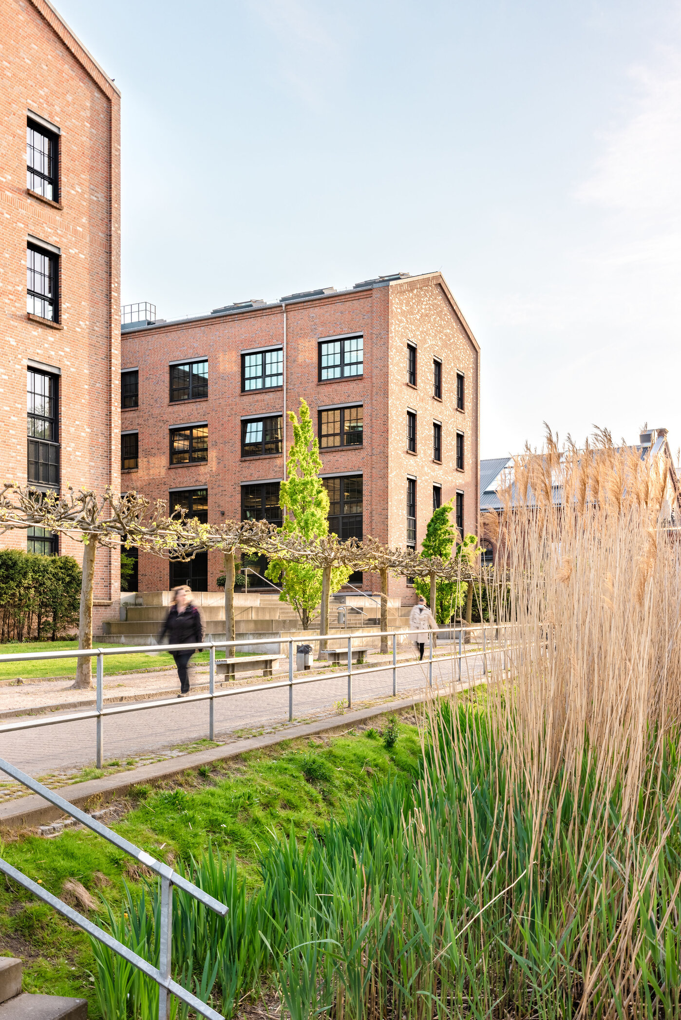 Urbanes Wohnambiente mit Natur. Ein modernes Backsteingebäude mit mehreren Fenstern steht im Vordergrund. Zwei Personen gehen auf einem Weg, der von Büschen und hohem Gras gesäumt ist. Im Hintergrund sind weitere Gebäude und ein klarer Himmel sichtbar.