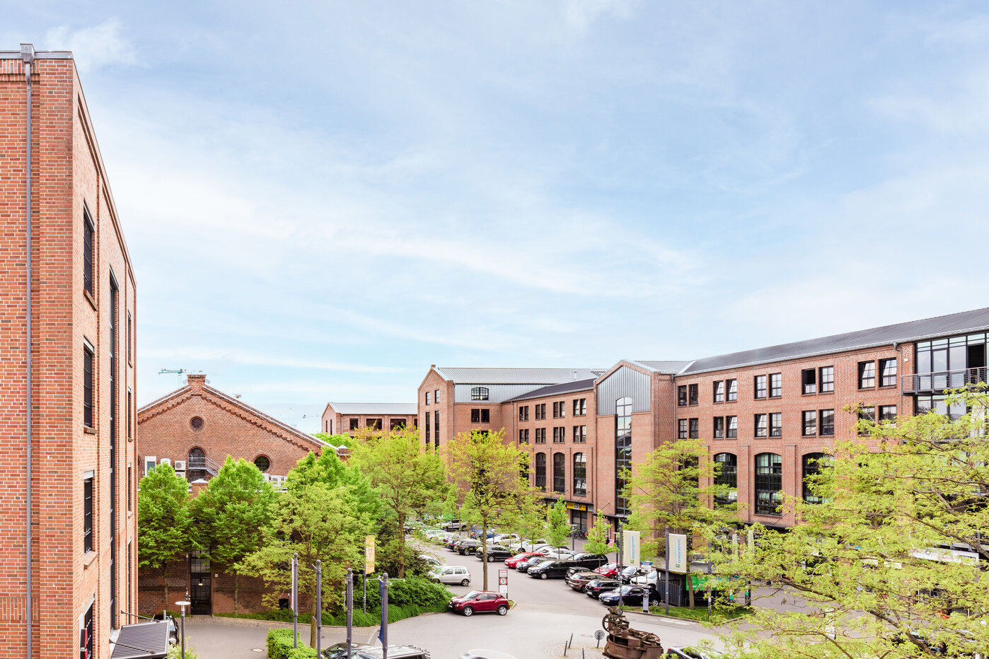 Städtische Architektur mit Grünflächen Eine Ansicht eines modernen Stadtviertels mit Backsteingebäuden und einer grünen Umgebung. Im Vordergrund sind parkende Autos und einige Bäume sichtbar, während der Himmel hell und klar ist. Die Architektur kombiniert historische und moderne Elemente.