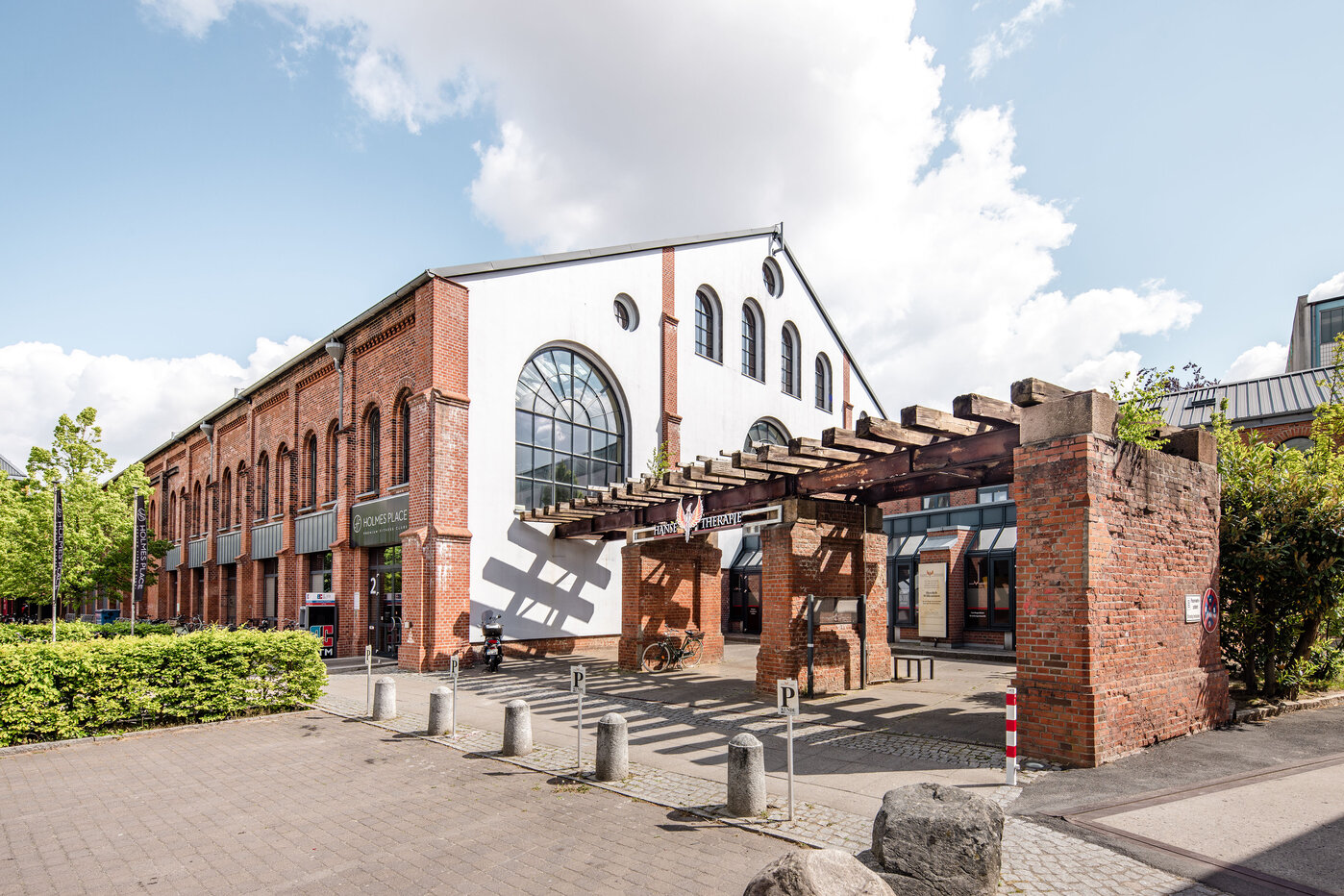 Ehemalige Fabrik im Stadtzentrum Ein historisches Backsteingebäude mit großen Fenstern und einem überdachten Eingangsbereich. Vor dem Gebäude steht eine Parkplatztabelle, und grüne Sträucher säumen den Weg. Es herrscht sonniges Wetter mit einigen Wolken am Himmel.