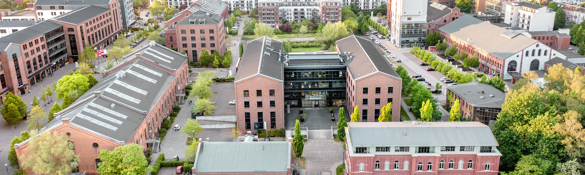 Luftaufnahme eines städtischen Gebäudekomplexes, umgeben von Bäumen und grünen Flächen. Die Architektur ist eine Mischung aus modernen und historischen Baustilen, mit mehreren Gebäuden und Parkplätzen. Der Hintergrund zeigt eine weitere städtische Umgebung mit Wohnungen und Gewerbe.