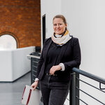 Eine Frau mit lässigem, professionellem Outfit steht an einer Treppe und lehnt sich leicht an ein Geländer. Sie hat einen strahlenden Ausdruck, hält eine Mappe in der Hand und trägt einen bunten Schal. Der Hintergrund zeigt eine moderne, helle Umgebung.