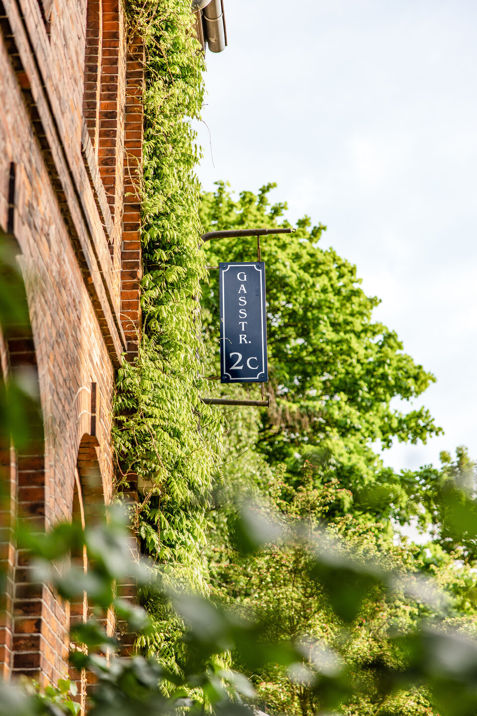 Ein braunrotes Gebäude ist mit grüner Vegetation bewachsen. An der Fassade hängt ein Schild mit der Aufschrift „Gästr. 2c“. Im Hintergrund sind Bäume und ein blauer Himmel zu sehen.