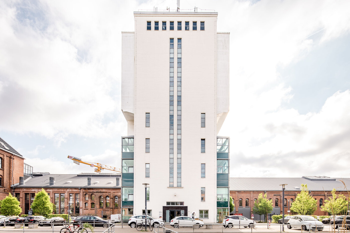 Moderne Architektur im Stadtzentrum Das Bild zeigt ein modernes, weißes Gebäude mit einer hohen, rechteckigen Form. Es hat große Glasfenster an der unteren Etage und eine markante vertikale Linie in der Mitte. Im Vordergrund stehen einige Autos und Bäume, die den städtischen Raum umrahmen.