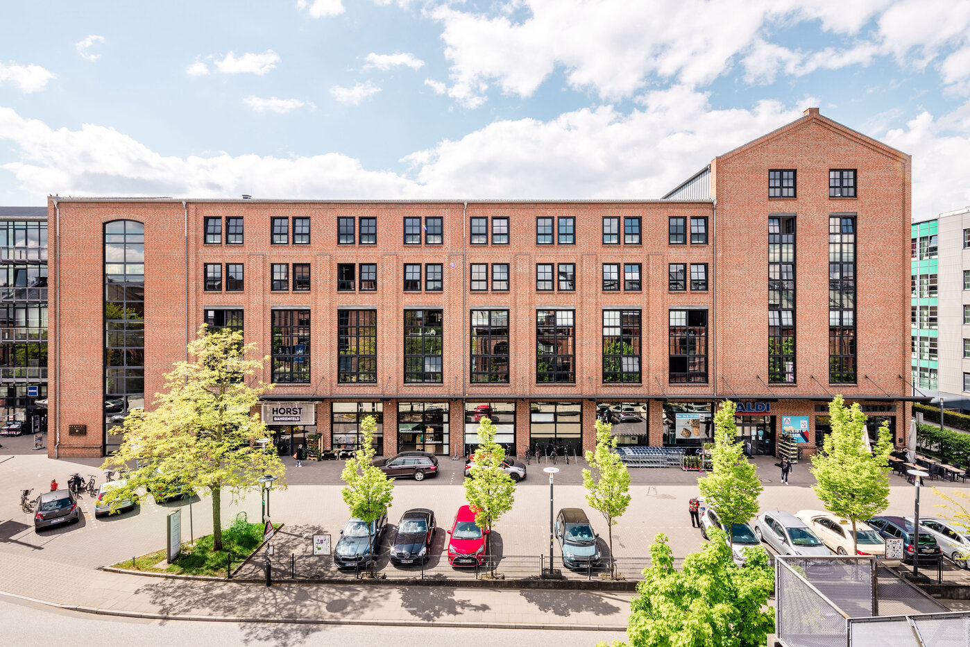 Modernes Backsteingebäude mit Bäumen Ein modernes, mehrstöckiges Gebäude aus roten Backsteinen mit großen Fenstern. Vor dem Haus stehen mehrere Bäume und Fahrzeuge. Die Umgebung wirkt urban und einladend, mit einem klaren Himmel und vereinzelten Wolken im Hintergrund.