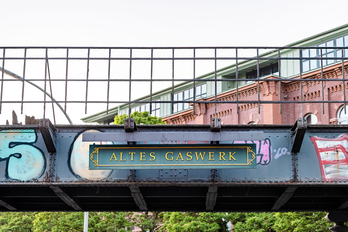 Altes Gaswerk Logo Ein schwarzes Metallbanner mit goldener Schrift trägt den Text „ALTES GASWERK“. Das Banner hängt über einer Brücke und ist umgeben von Graffiti. Im Hintergrund sind moderne Gebäude und grüne Bäume sichtbar.