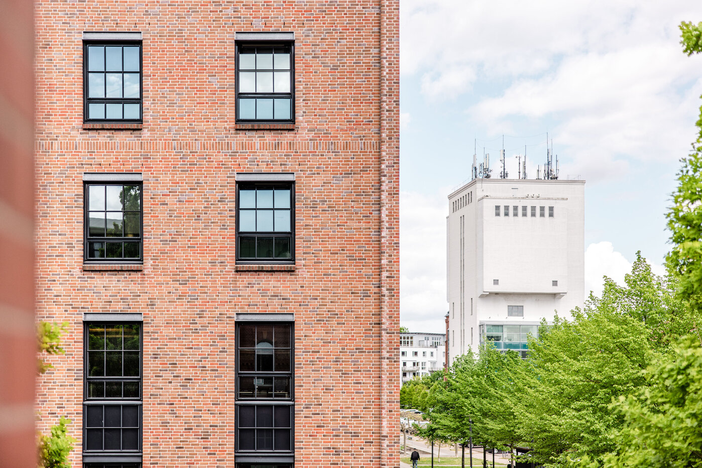 Städtische Architektur mit Grüner Umgebung Zwei Gebäude sind im Vordergrund sichtbar: ein Backsteingebäude mit großen, schwarzen Fenstern und ein modernes, helles Gebäude im Hintergrund. Dazwischen sind grüne Bäume zu sehen, die eine ruhige Umgebung schaffen. Der Himmel ist leicht bewölkt.