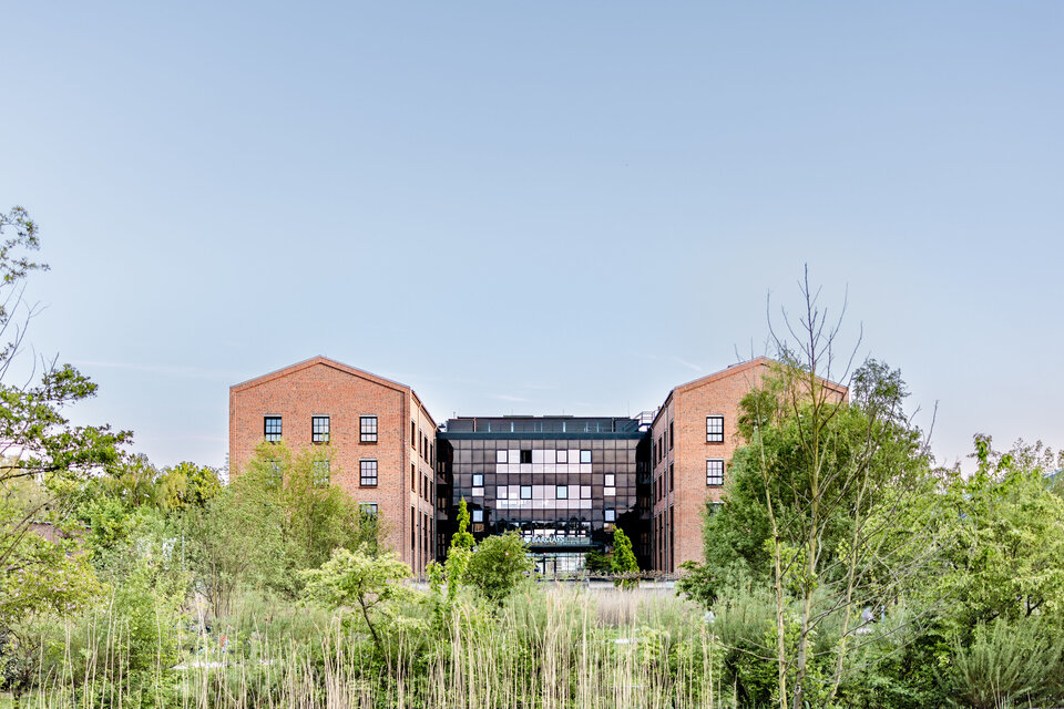Ein modernes Gebäude mit einer Glasfassade und roten Backsteinen, umgeben von grünen Bäumen und Sträuchern. Die Architektur kombiniert klassische und moderne Elemente und spiegelt eine naturnahe Gestaltung wider.