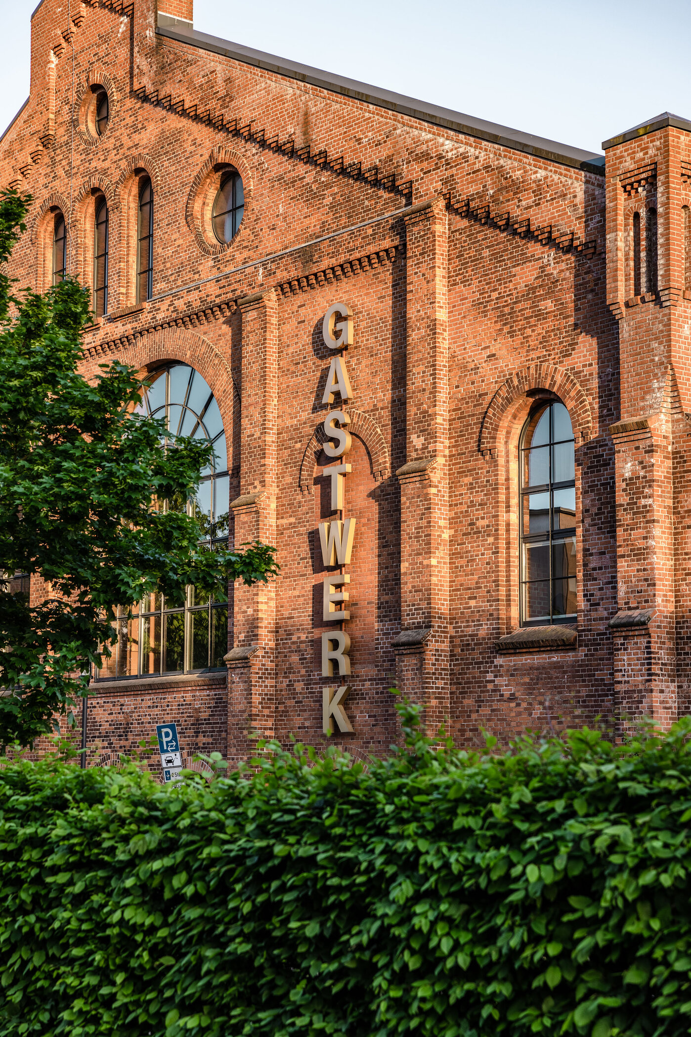 Historisches Gaswerk-Gebäude Ein historisches Gebäude aus roten Ziegeln mit der Aufschrift „GASWERK“. Die Fassade zeigt große Fenster und ist von grünen Sträuchern umgeben. Das Bild vermittelt ein Gefühl von industriellem Charme und Architektur.