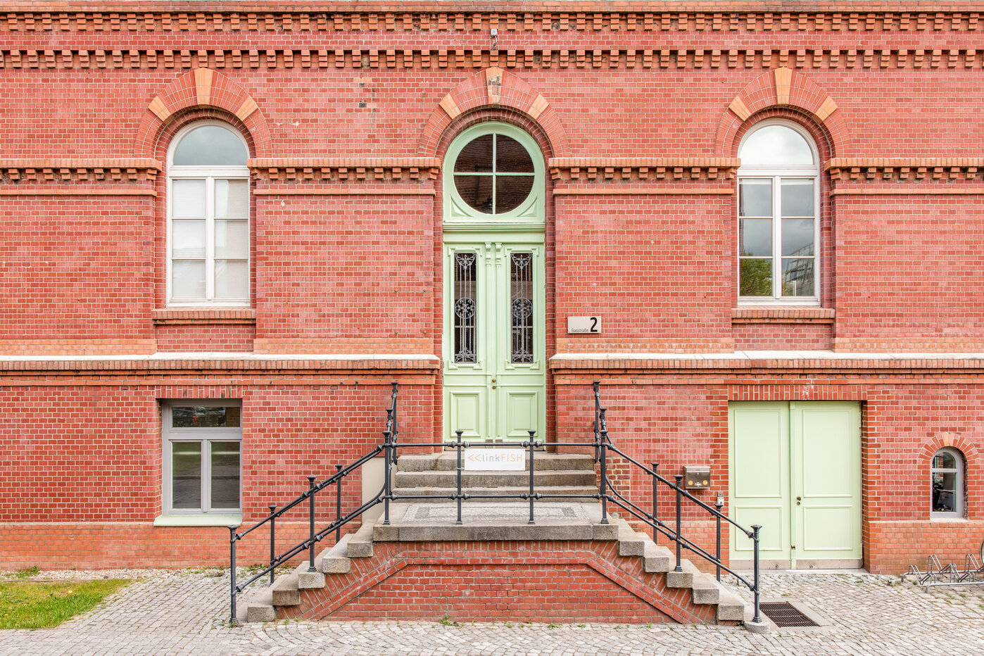 Historisches Ziegelgebäude mit Eingang Eine rote Backsteinfassade mit einem großen, grünen Eingangstür und runden Fenstern. Eine Zugangsrampe führt zu den Stufen vor der Tür. Neben der Eingangstür befindet sich eine kleinere, grüne Tür. Der Bereich ist von einem gepflasterten Gehweg umgeben.