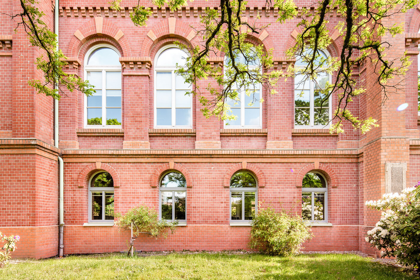 Historische Ziegelarchitektur mit Fenstern Eine rote Backsteinmauer mit großen, hohen Fenstern, die in verschiedenen Bögen angeordnet sind. Die Fenster sind von grünen Bäumen und Sträuchern umgeben. Das Bild vermittelt eine ruhige und harmonische Atmosphäre.