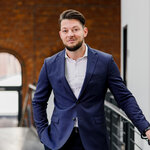 Ein Mann mit kurzen, dunklen Haaren steht in einem modernen Raum mit Ziegelwänden. Er trägt einen blauen Anzug mit weißem Hemd und lächelt. Im Hintergrund ist ein Geländer und ein Fenster sichtbar, was dem Bild eine professionelle Atmosphäre verleiht.