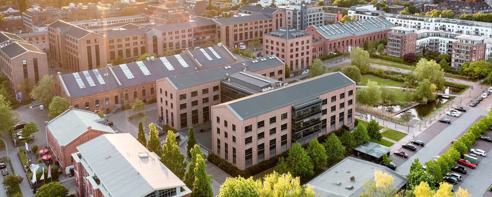 Luftaufnahme moderner Stadtarchitektur Luftaufnahme eines städtischen Gebiets mit modernen Gebäuden, umgeben von Bäumen und Grünflächen. Die Sonne scheint über der Szene und wirft warmes Licht auf die Architektur. Ein ruhiger Wasserlauf verläuft durch das Gebiet und es sind Parkplätze sichtbar.
